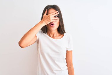 Genç ve güzel bir kadın, günlük tişört giyip izole edilmiş beyaz arka planda dikiliyor. Şoka giriyor, yüzlerini ve gözlerini eliyle kapatıyor ve utanmış bir ifadeyle parmaklarından bakıyor..