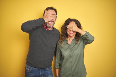 Güzel bir orta yaşlı çift, izole edilmiş sarı arka planda gülümsüyor ve elleri yüzlerinde şaşkınlıktan gözleri kapatıyordu. Kör kavram.