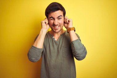 Gürültülü müzik sesi için sarı arka planda dikilen ve kulaklarını parmaklarıyla kapatan sıradan tişört giyen genç yakışıklı bir adam. Sağır kavramı.