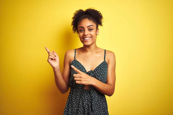 African american woman wearing summer casual green dress over isolated yellow background smiling and looking at the camera pointing with two hands and fingers to the side.