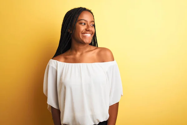 Young african american woman wearing white t-shirt standing over isolated yellow background looking away to side with smile on face, natural expression. Laughing confident.