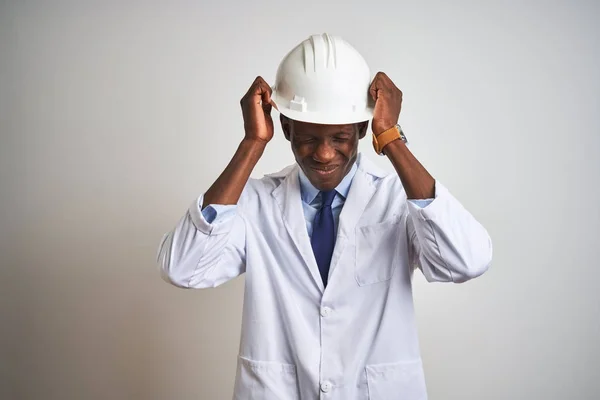 African american engineer man wearing coat and helmet over isolated white background suffering from headache desperate and stressed because pain and migraine. Hands on head.