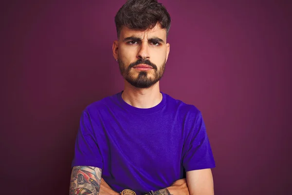 Young man with tattoo wearing t-shirt standing over isolated purple background skeptic and nervous, disapproving expression on face with crossed arms. Negative person.