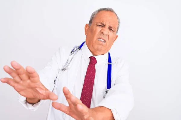 Senior grey-haired doctor man wearing stethoscope standing over isolated white background disgusted expression, displeased and fearful doing disgust face because aversion reaction. With hands raised. Annoying concept.