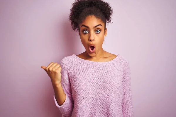 Young african american woman wearing winter sweater standing over isolated pink background Surprised pointing with hand finger to the side, open mouth amazed expression.