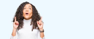 Kıvırcık saçlı Genç güzel kadın balıkçı yaka kazak şaşırmış ve görünümlü ve parmaklar ve yükseltilmiş kollar ile işaret sürpriz giyiyor.