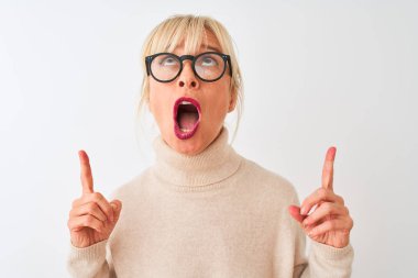 Balıkçı yaka kazak ve gözlük takan orta yaşlı bir kadın izole edilmiş beyaz arka planda şaşkın şaşkın bakıyor, parmaklarıyla işaret ediyor ve kollarını kaldırıyordu..