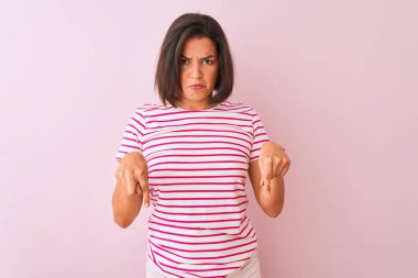 İzole edilmiş pembe arka plan üzerinde duran çizgili tişört giyen genç ve güzel bir kadın üzgün ve üzgün görünüyor, parmaklarıyla yönü gösteriyor, mutsuz ve depresif..