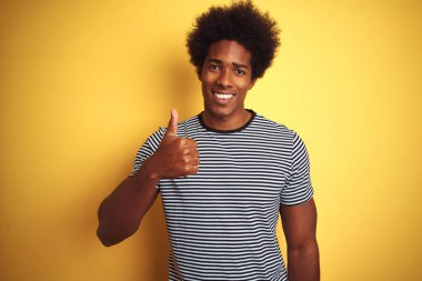 Afro saçlı, donanma çizgili tişörtlü, izole edilmiş sarı arka planda mutlu bir el hareketi yapan Afro-Amerikan bir adam. Kameraya bakıp başarıyı gösteren ifade onaylanıyor.