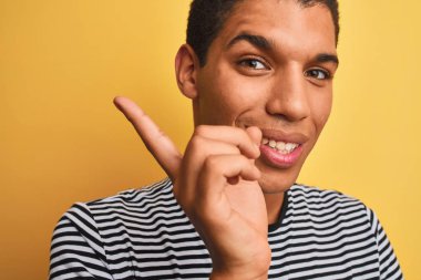 İzole edilmiş sarı arka plan üzerinde denizci çizgili tişört giyen genç yakışıklı Arap adam elini ve parmağını kenarı göstererek çok mutlu oluyor.