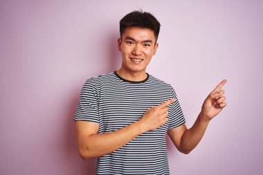 Genç Asyalı, çizgili tişört giyen, pembe arka planda duran, gülümseyen ve kameraya bakan, iki eliyle ve parmaklarıyla yüzünü gösteren Asyalı bir adam..