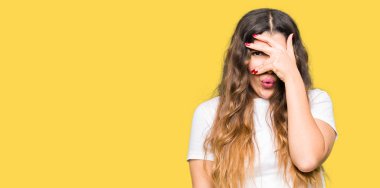 Mahcup ifadeyle parmaklarımın arasından seyir yüz ve gözler el ile kapsayan şok bakmak rahat beyaz t-shirt giyen genç güzel kadın.