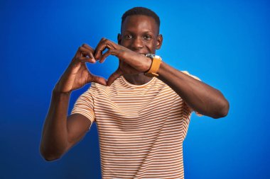 Siyah çizgili günlük tişört giyen Afro-Amerikan bir adam izole edilmiş mavi arka planda duruyor. Aşkla gülümsüyor ve elleriyle kalp sembolü ve şekli gösteriyor. Romantik konsept.