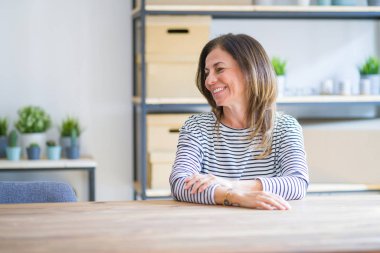 Orta yaş yaşlı kadın evde masada oturan yüzünde gülümseme ile yan bakıyor, doğal ifade. Gülmekten emin.