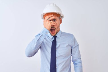 Young business man wearing contractor safety helmet over isolated background peeking in shock covering face and eyes with hand, looking through fingers with embarrassed expression.