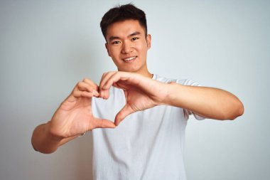 Asyalı genç bir Çinli tişört giyiyor. İzole edilmiş beyaz arka planda duruyor. Aşkla gülümsüyor, elleriyle kalp sembolü yapıyor. Romantik konsept.