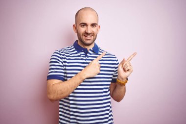 Sakallı genç ve kel bir adam pembe izole edilmiş arka planda günlük mavi tişört giyiyor gülümsüyor ve kameraya bakıyor iki eliyle ve parmaklarıyla yüzünü gösteriyor..