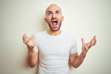 Sakallı genç ve kel bir adam izole edilmiş arka planda günlük beyaz tişört giyiyor çılgın ve sinirli bir şekilde bağırıyor ve agresif bir ifade ve kollar havada bağırıyor. Hayal kırıklığı konsepti.