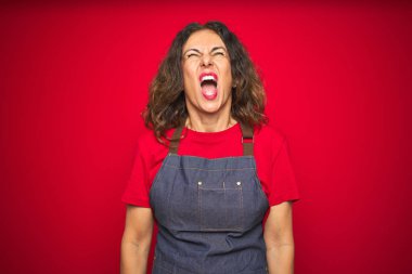 Orta yaş yaşlı kadın kırmızı izole arka plan üzerinde önlük üniforma giyen kızgın ve deli çığlık sinirli ve öfkeli, öfke ile bağırarak. Öfke ve agresif kavram.