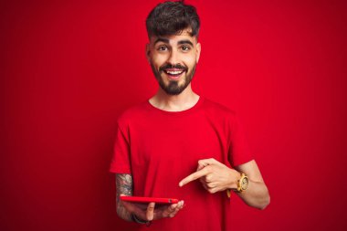 Dövmeli genç adam, izole edilmiş kırmızı arka planda duran tablet kullanıyor. Elle ve parmakla işaret ederek çok mutlu.