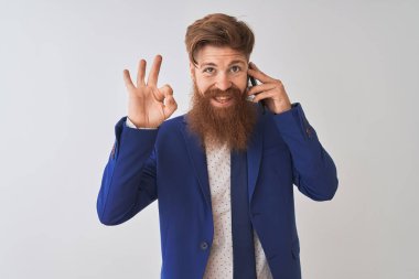 Genç kızıl saçlı İrlandalı işadamı, izole edilmiş beyaz arka plan üzerinden akıllı telefondan konuşuyor. Tamam işareti yapıyor, mükemmel bir sembol.
