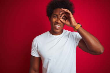 Afro saçlı, beyaz tişörtlü, izole edilmiş kırmızı arka planda dikilen genç bir Amerikalı. El hareketleriyle gülümsüyor, gözleriyle mutlu yüzüyle parmaklarının arasından bakıyor..