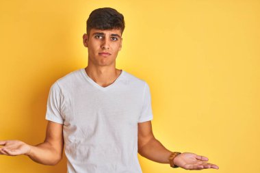 İzole edilmiş sarı arka planda duran beyaz tişörtlü genç Hintli adam hiçbir şey bilmiyor ve kolları açık, hiçbir fikri yok..