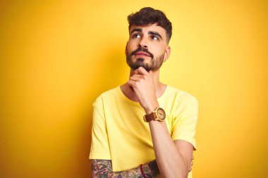 Dövmeli, tişörtlü genç adam izole edilmiş sarı arka planda duruyor, elleri çenesinde soru sorarken, düşünceli bir ifade takınıyor. Düşünceli bir yüzle gülümsüyor. Şüphe kavramı.