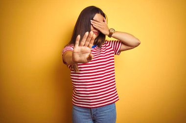 Çizgili tişört giyen genç ve güzel bir kadın izole edilmiş sarı arka plan üzerinde duruyor gözleri ellerle örtüyor ve üzgün ve korku ifadeleriyle jest yapıyor. Utanç verici ve olumsuz bir kavram..