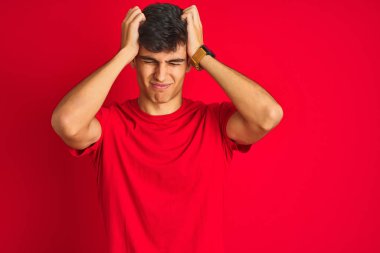 Genç Hintli adam, izole edilmiş kırmızı arka plan üzerinde dikilip acı ve migren yüzünden çaresiz ve stresli bir baş ağrısı çeken bir tişört giyiyor. Ellerini başının üstüne koy..
