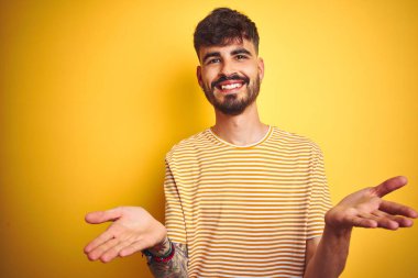 Dövmeli, çizgili tişörtlü genç adam izole edilmiş sarı arka planda duruyor. Gülümsüyor, yardım eli uzatıyor ve kabul ediyor..