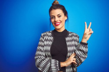 Mavi izole edilmiş arka planda duran ceketini giymiş genç ve güzel bir kadın kameraya bakıp zafer işareti yaparken gülümsüyor. İki numara..
