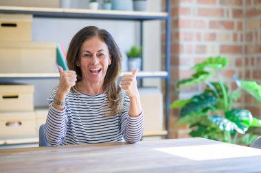 Orta yaş yaşlı kadın evde masada oturan başarı işareti el ile olumlu jest yapıyor, gülümseyerek ve mutlu başparmak kadar. Neşeli ifade ve kazanan jest.