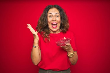 Orta yaşlı bir kadın kırmızı izole arka planda ahududu yiyor çok mutlu ve heyecanlı, kazanan yüz ifadesi zaferi büyük bir gülümsemeyle kutluyor ve ellerini kaldırıyor.