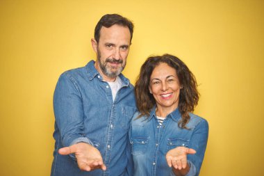 Güzel bir orta yaşlı çift, izole edilmiş sarı arka planda durup gülümsüyor, avuç içi kadar yardım ve kabul görüyor..