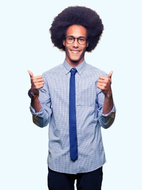 El ile pozitif jest yapıyor gözlük başarı işareti giyiyor afro saçlı genç Afro-Amerikan iş adamı başparmak gülümseyen ve mutlu. Neşeli bir ifadeyle, kazanan jest kameraya bakıyor.