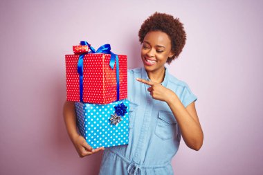 Afrika kökenli Amerikalı bir kadın pembe izole edilmiş arka planda doğum günü hediyelerini tutarken mutlu bir şekilde işaret ediyor.