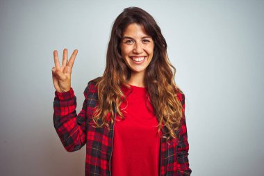 Kırmızı tişört ve ceket giyen genç güzel bir kadın beyaz izole edilmiş arka plan üzerinde duruyor ve üç numaralı parmaklarıyla işaret ederken kendinden emin ve mutlu bir şekilde gülümsüyor..