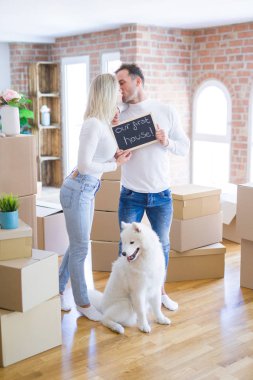 Karton kutular etrafında yeni evde öpüşme mesajı ile tahta tutan ayakta köpek ayakta genç güzel çift