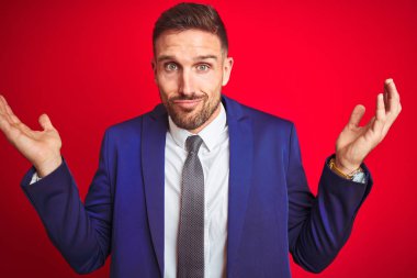 Genç ve yakışıklı iş adamı, kırmızı arka plan üzerinde soyutlanmış kolları ve elleri havada, kafası karışık bir yüz ifadesi. Şüphe kavramı.