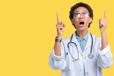 İzole arka plan üzerinde tıbbi ceket giyen genç Afro-Amerikan doktor kadın hayrete ve yukarıya bakıyor ve parmakları ile işaret ve silah kaldırdı şaşırttı.