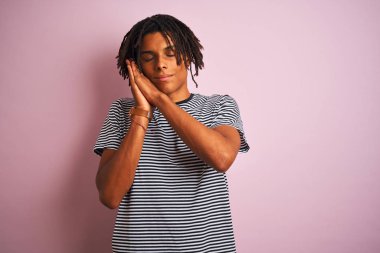 Sarı çizgili tişört giyen Afro adam izole edilmiş pembe arka planda ayakta uyur, yorgun düşler kurar ve elleri ve elleri kapalı gülümserken birlikte poz verir..