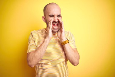Sakallı, sarı tişörtlü, sakallı genç bir adam. İzole edilmiş sarı arka plan. Elleri ağzında kızgın bir şekilde bağırıyor.