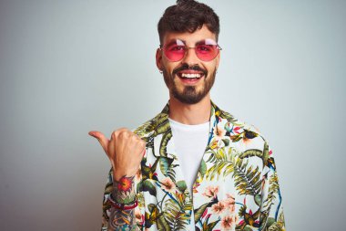 Tatilde dövmeli, güneş gözlüklü, izole edilmiş beyaz arka planda gülümseyen, gülümseyen ve baş parmağıyla yüzünü gösteren bir adam..