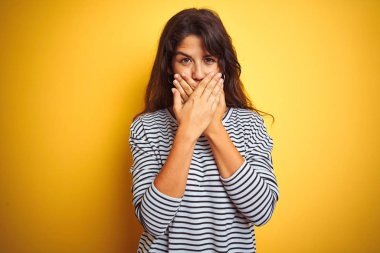 Sarı yalıtılmış arka plan üzerinde duran çizgili tişört giyen genç ve güzel bir kadın yanlışlıkla ağızlarını kapatarak şok geçirdi. Gizli konsept.