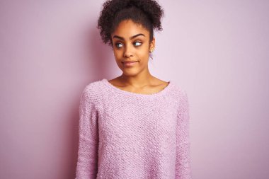 Kış süveteri giyen genç Afrikalı Amerikalı kadın izole edilmiş pembe arka planda duruyor gülümsüyor ve başka tarafa bakıyor..