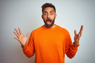 Young indian man wearing orange sweater over isolated white background celebrating crazy and amazed for success with arms raised and open eyes screaming excited. Winner concept