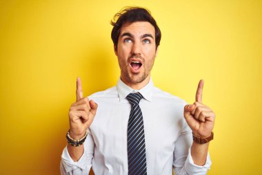 Zarif gömlek ve kravat giyen genç yakışıklı iş adamı, izole edilmiş sarı arka planda şaşkın şaşkın ve şaşırmış bir şekilde yukarı bakıyor ve parmaklarıyla işaret ediyor ve kollarını kaldırıyordu..