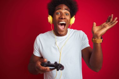 Oyun oynayan Afro Oyuncu video oyunu oynayan kırmızı arka plan üzerinde joystick kulaklıklar kullanıyor çok mutlu ve heyecanlı, kazanan yüz ifadesi zaferi büyük bir gülümsemeyle kutluyor ve ellerini kaldırıyor.