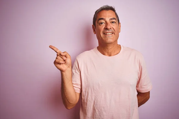 stock image Handsome middle age man wearing t-shirt standing over isolated pink background with a big smile on face, pointing with hand and finger to the side looking at the camera.